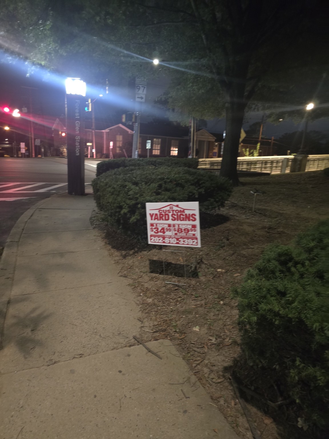 Red and white BoldStake custom yard sign displayed on a metal stake beside a sidewalk near Forest Glen Station at night.