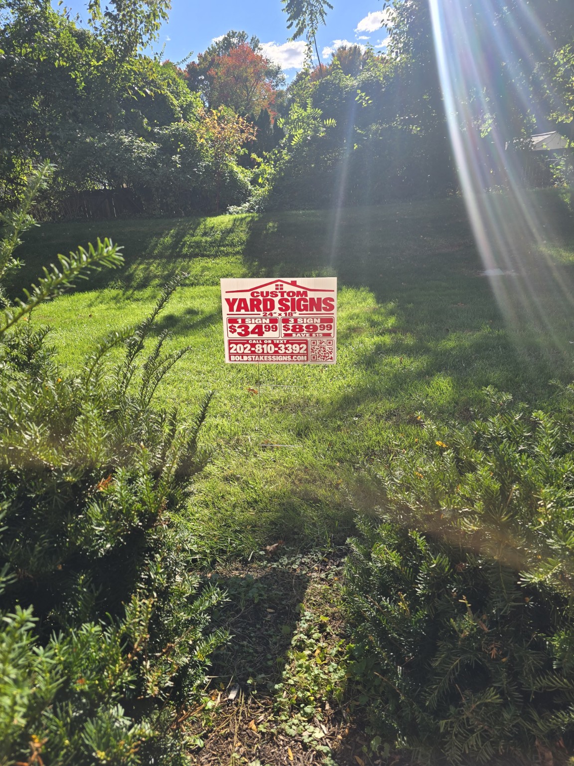 Red and white Bold Stakes custom yard sign displayed on a metal H-stake in a sunny green lawn surrounded by trees.